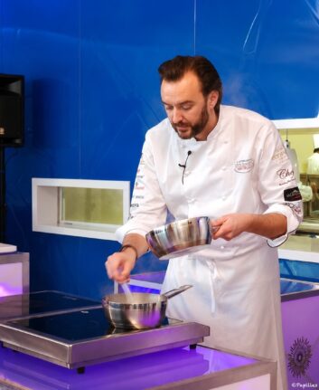 Chef in a white coat stirs a sauce in a stainless steel pot on an induction cooktop during a cooking demonstration, blue backdrop behind him.